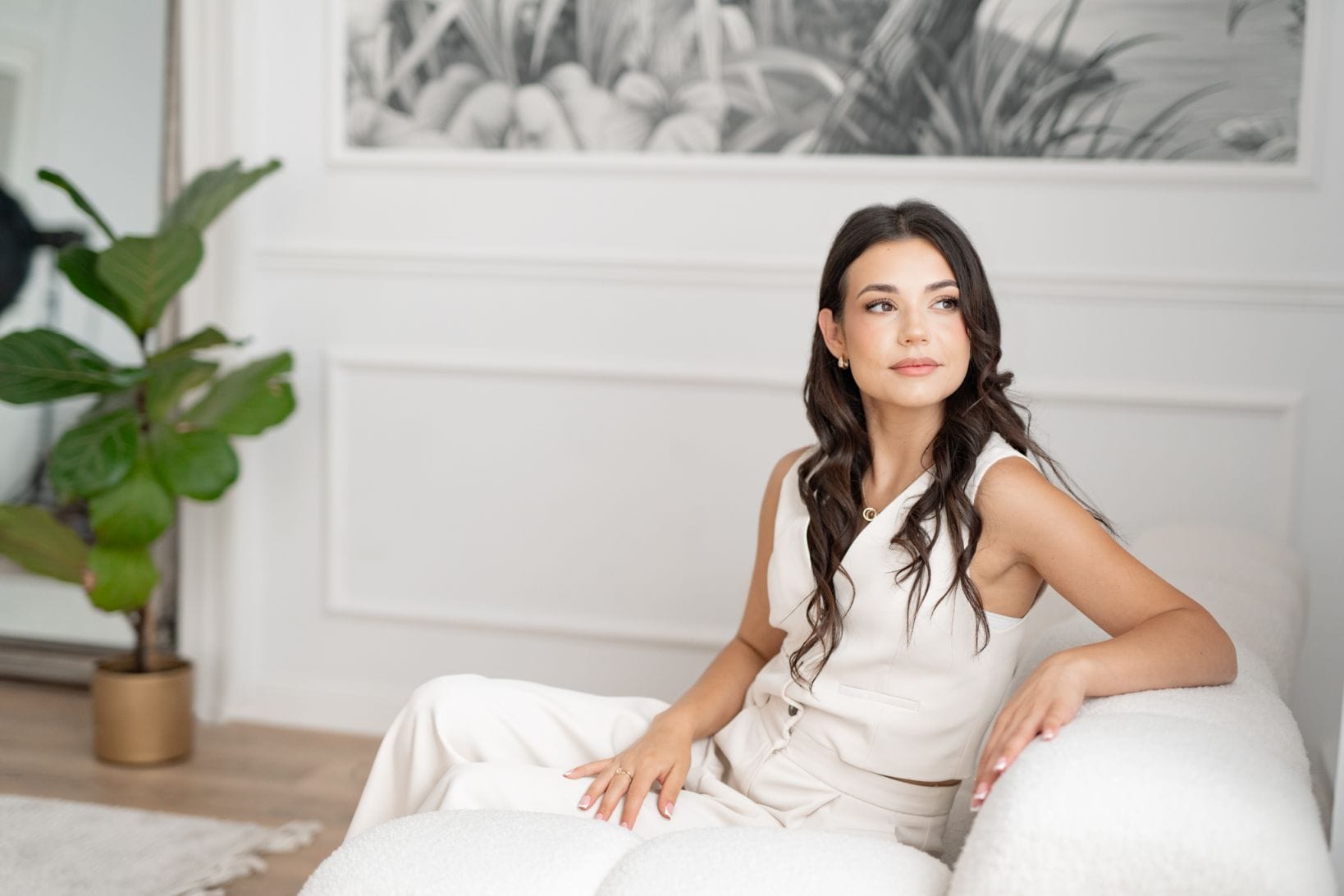 Kobieta o długich ciemnych włosach, ubrana w biały strój bez rękawów, siedzi na białym tapicerowanym krześle w jasnym, nowoczesnym pokoju. Za nią znajduje się duża roślina w złotej doniczce i monochromatyczna grafika z motywem natury na ścianie. W zamyśleniu spogląda w bok, chwytając inspirujący moment na zdjęcia dla szkół językowych - pojawiłaca się sesja fotograficzna.
