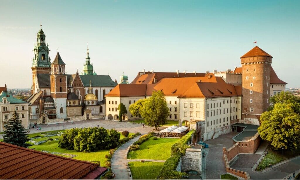 Panoramiczny widok Zamku Wawelskiego w Krakowie, w Polsce. Zabytkowy kompleks łączy w sobie wiele stylów architektonicznych, otoczony zielonymi trawnikami i kamiennymi ścieżkami. Wieże i iglice zamku wznoszą się na tle czystego, błękitnego nieba. Stanowi tło dla rankingu fotografów w Krakowie.