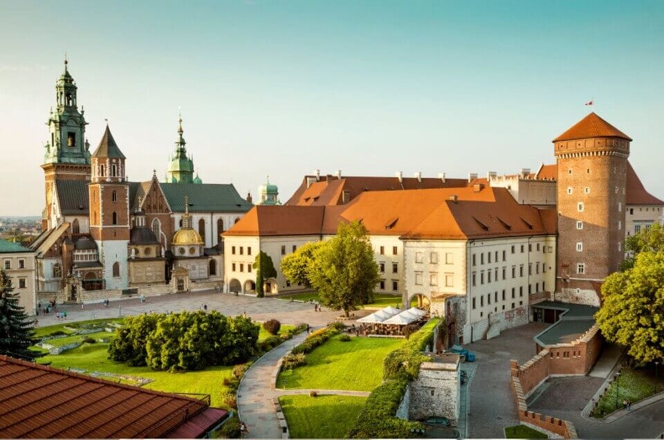 Panoramiczny widok Zamku Wawelskiego w Krakowie, w Polsce. Zabytkowy kompleks łączy w sobie wiele stylów architektonicznych, otoczony zielonymi trawnikami i kamiennymi ścieżkami. Wieże i iglice zamku wznoszą się na tle czystego, błękitnego nieba. Stanowi tło dla rankingu fotografów w Krakowie.